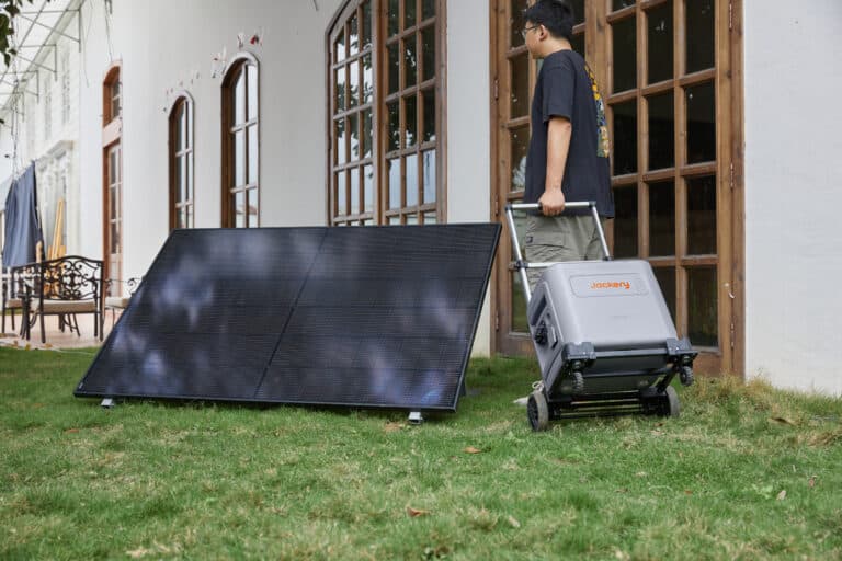 Flexibles Solarmodul mit mobilen Heimspeicher auf Trolley in der Hand einer männlichen Person vor einem Haus